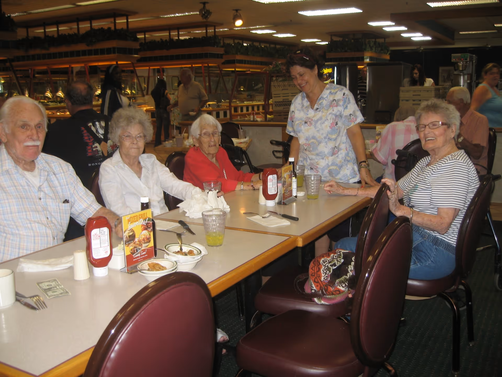 A group of elderly people sitting around a dining table in a cafeteria-style setting, with a caregiver standing and smiling beside them. The table has condiments, glasses with drinks, and some plates with food. Other people and buffet counters are visible in the background.