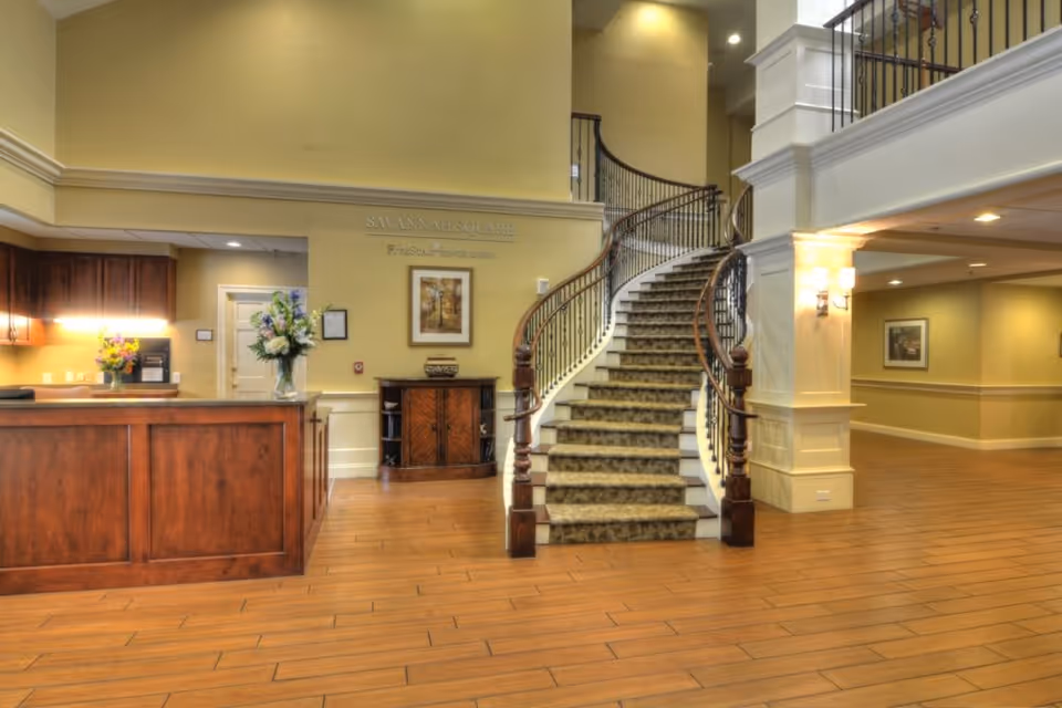 Interior view of Savannah Square facility featuring a wooden reception desk with flower arrangements, a carpeted curved staircase with wooden handrails, warm yellow walls, wooden flooring, and soft lighting fixtures.