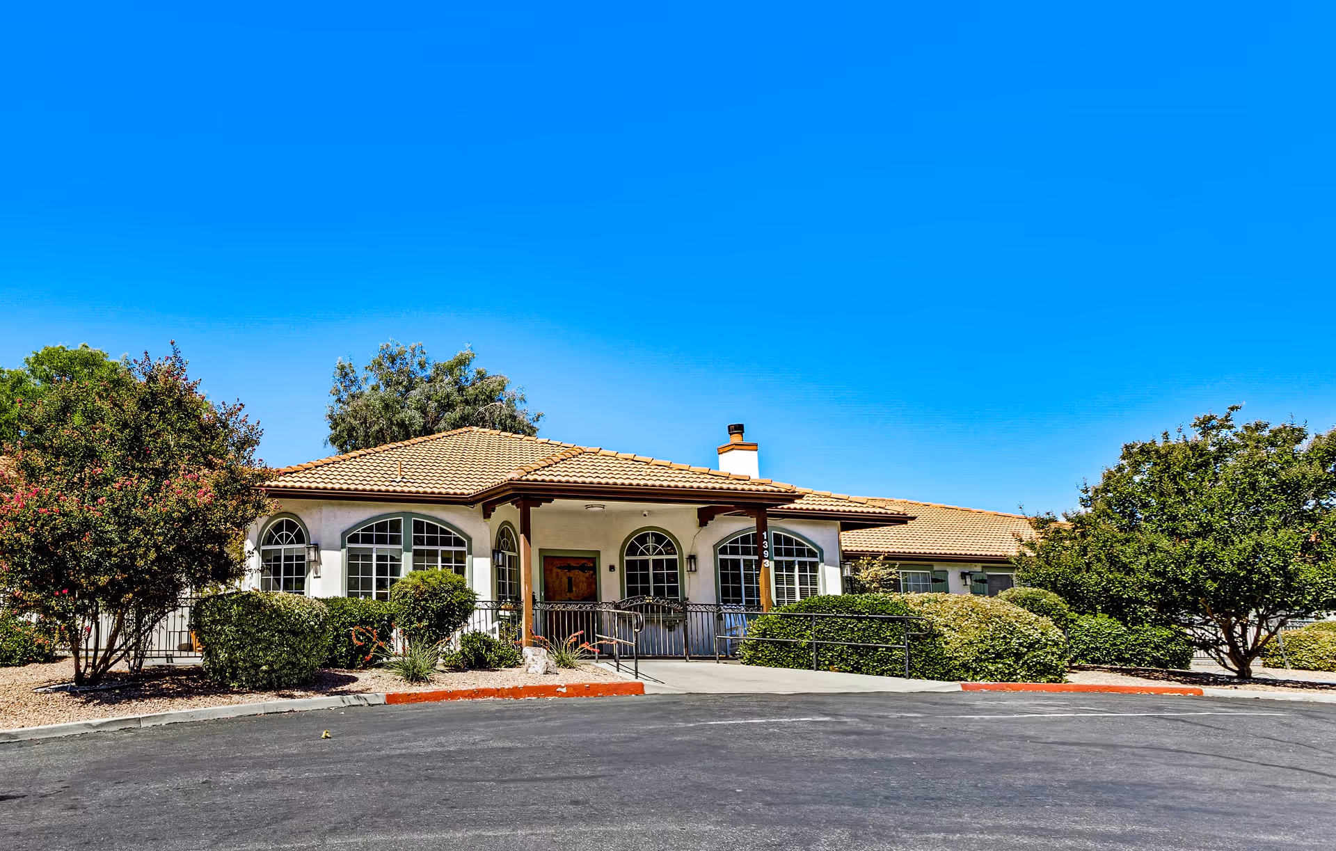 Single-story assisted living building with a tiled roof, arched windows, a central entrance ramp and landscaped shrubs under a clear blue sky.