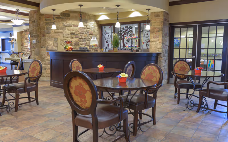 A cozy dining area with round wooden tables and cushioned chairs featuring floral upholstery. The room has a tiled floor, stone accent walls, and a counter with hanging pendant lights. Small flower pots are placed on each table, and there are glass doors leading to another room in the background.