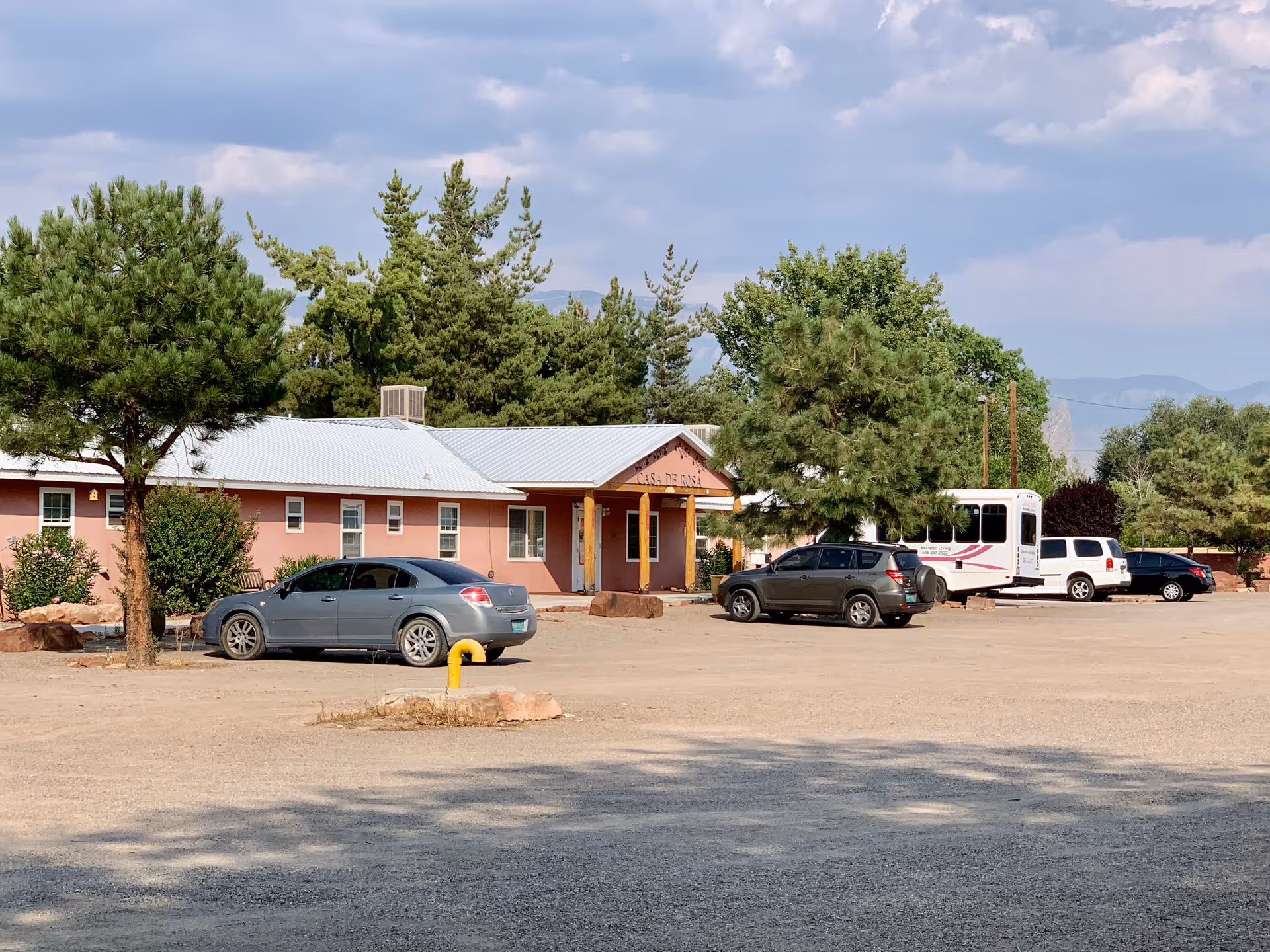 Exterior view of Casa de Rosa Assisted Living facility showing a single-story building with a metal roof, surrounded by trees and several parked cars in a gravel parking lot under a partly cloudy sky.