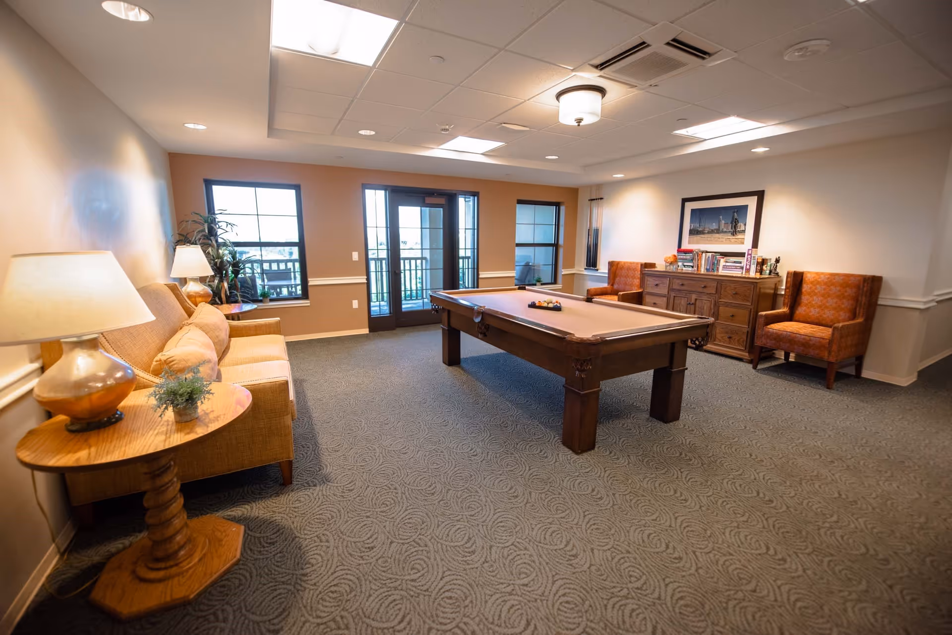 A well-lit recreational room with a pool table in the center, a beige sofa with cushions and a side table with a lamp and plant on the left, two orange patterned armchairs and a wooden dresser with books and decorative items on the right, and large windows and a glass door leading to an outdoor balcony.