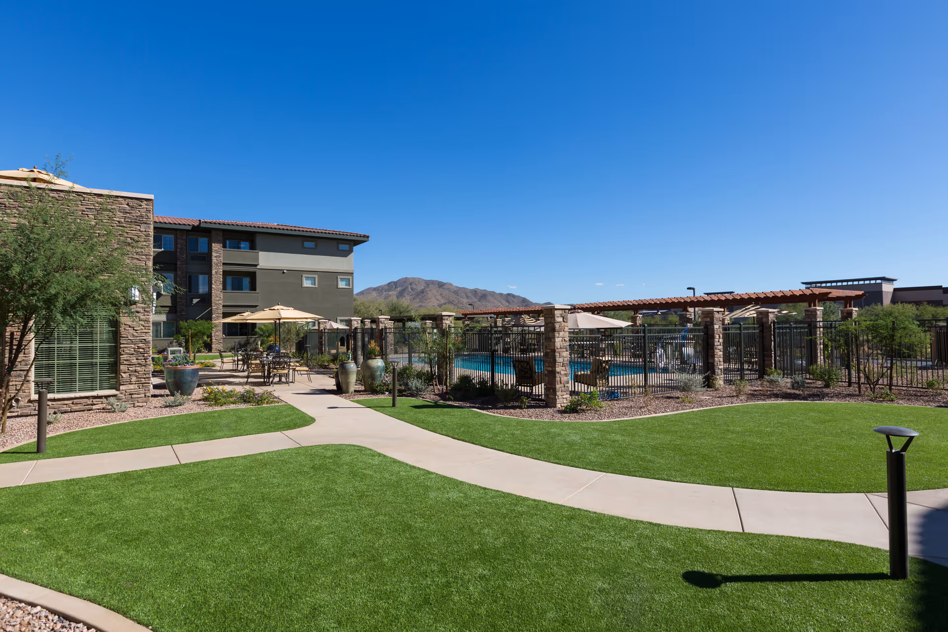 Outdoor area of The Enclave at Anthem Senior Living featuring a well-maintained lawn, paved walkways, patio tables with umbrellas, a fenced swimming pool, and a multi-story building with stone and stucco exterior under a clear blue sky with mountains in the background.