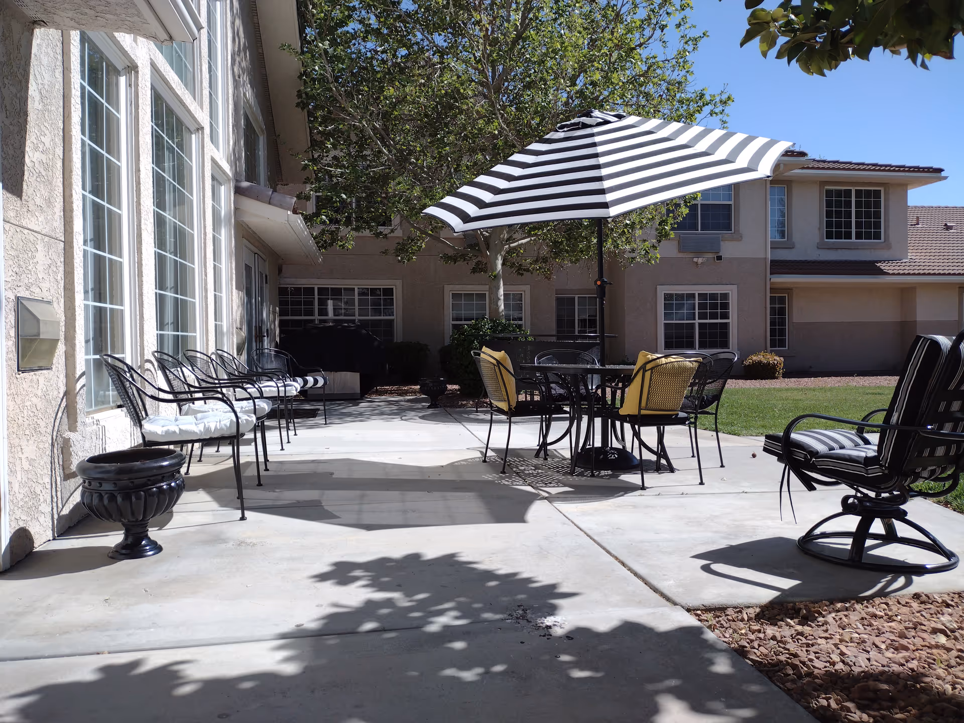 Outdoor patio area at Bellamar Lancaster with metal chairs lined up against the building wall, a round table with a black and white striped umbrella, and cushioned chairs on a concrete surface. There is a tree providing shade and a grassy area in the background.