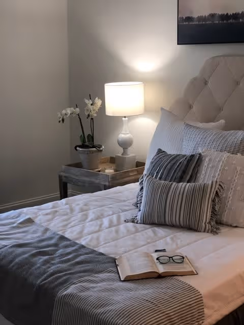 A cozy bedroom featuring a neatly made bed with multiple decorative pillows and a striped throw blanket. On the bed lies an open book and a pair of eyeglasses. Next to the bed is a wooden nightstand with a white table lamp and a potted orchid. The wall behind the bed is light-colored and has a framed picture hanging above the headboard.