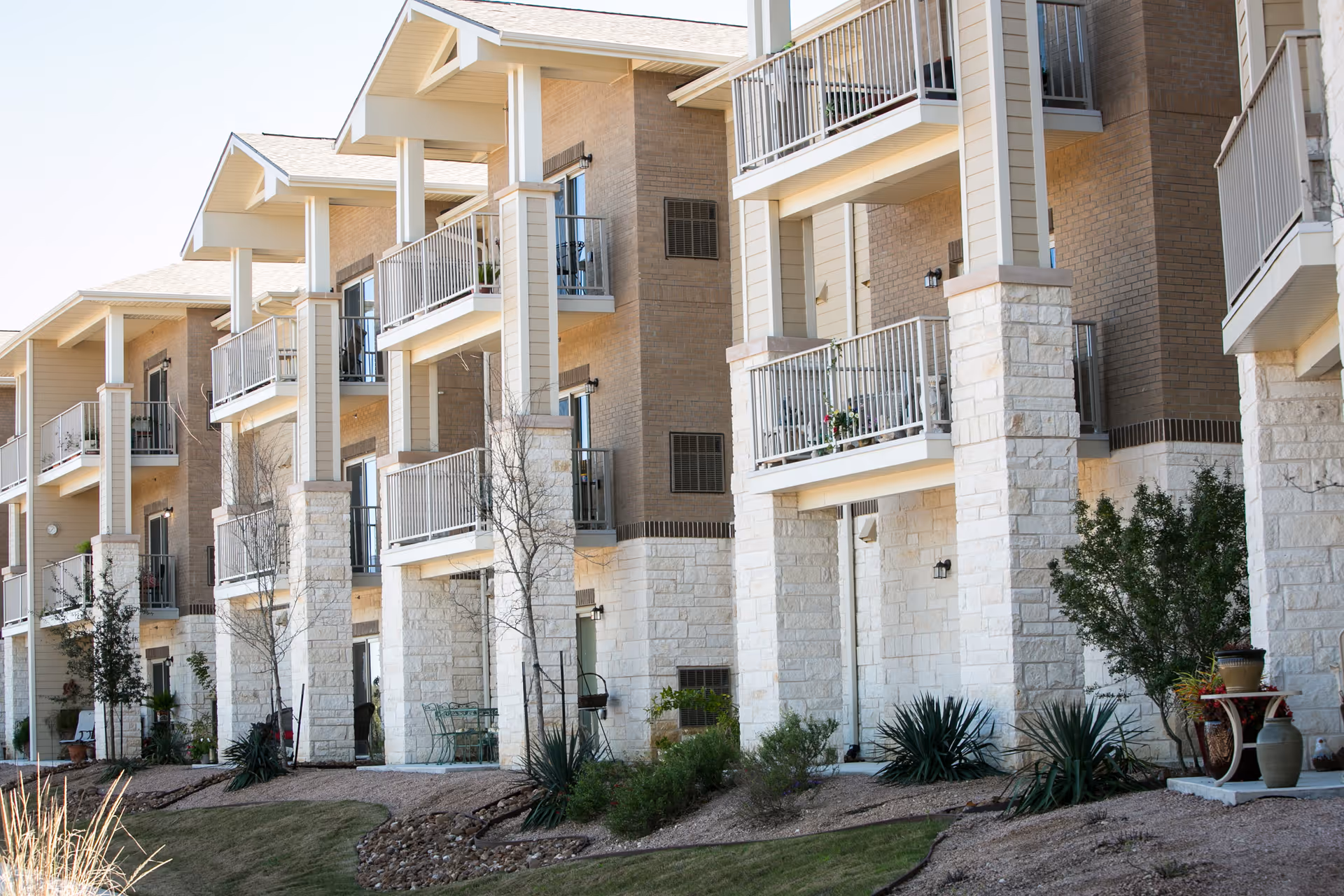 Exterior view of a multi-story residential building with balconies and patios, surrounded by landscaped greenery and small trees.