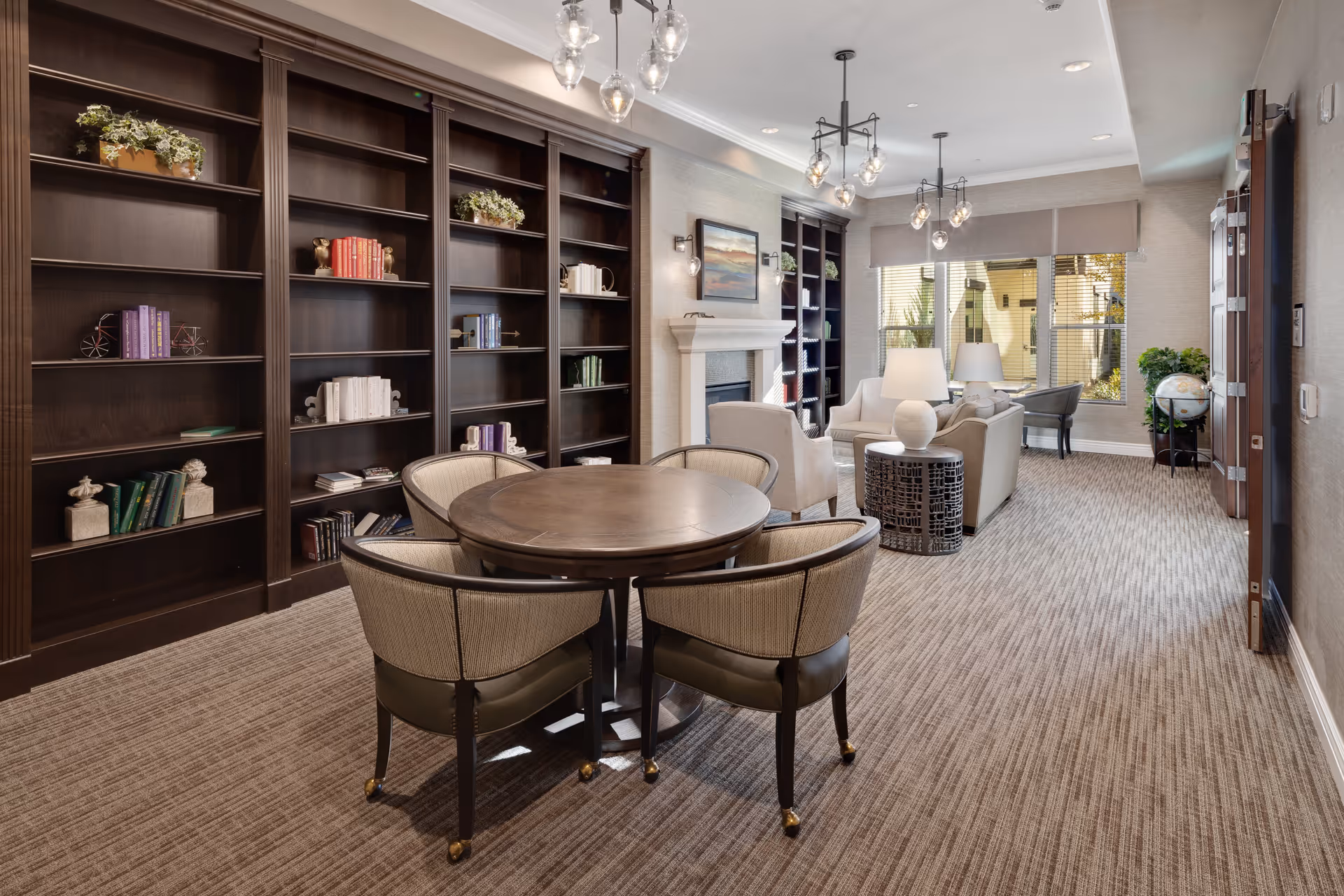 A cozy and well-lit common area featuring a round wooden table with four upholstered chairs in the foreground. Behind the table, there are tall dark wooden bookshelves with various books and decorative items. The room also includes a seating area with armchairs, a sofa, side tables with lamps, a fireplace, and large windows with blinds allowing natural light to enter.