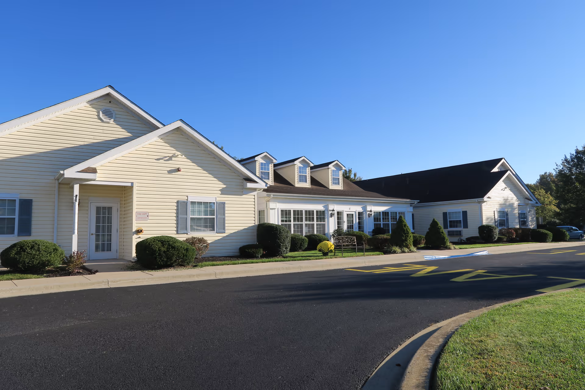 Front exterior of a single-story light-yellow assisted living building with shrubs, windows, and a paved driveway under a clear blue sky.