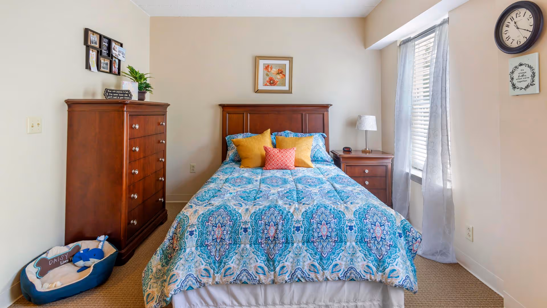Sunlit bedroom with a wooden bed, matching dresser and nightstand, a blue patterned comforter and decorative pillows, and a window with curtains.