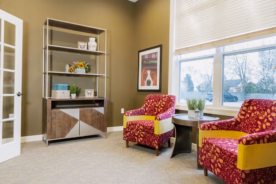 A cozy sitting area with two red and yellow patterned armchairs separated by a small round wooden table with a plant on it. Behind the chairs is a large window with blinds partially open, letting in natural light. To the left, there is a modern shelving unit with decorative items and a framed picture of a dog on the wall. The room has beige carpet and olive green walls.