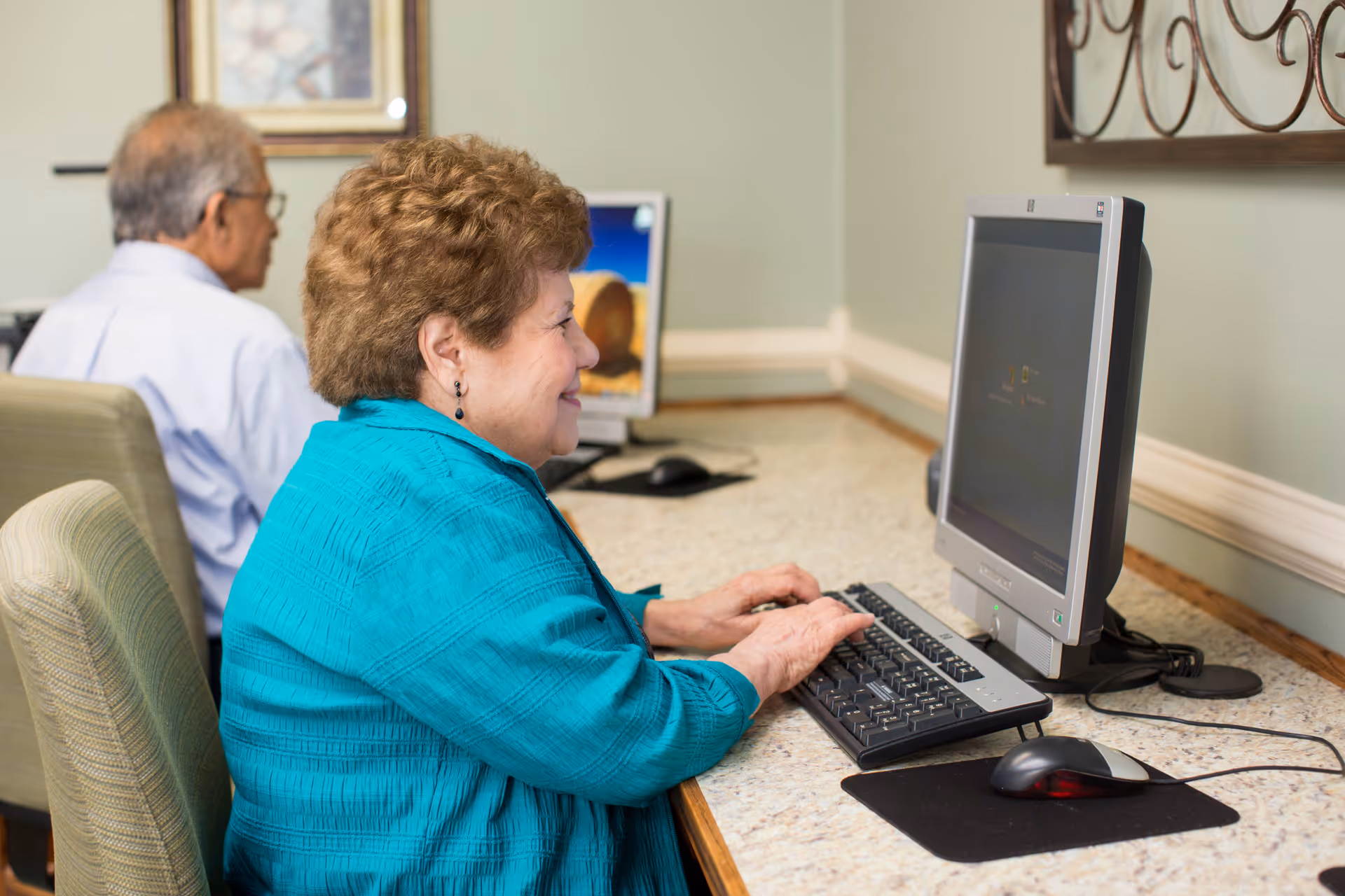 Two elderly individuals sitting side by side at a computer station in a room, with one woman in a turquoise jacket smiling and typing on a keyboard, and a man in a light blue shirt using a computer in the background.