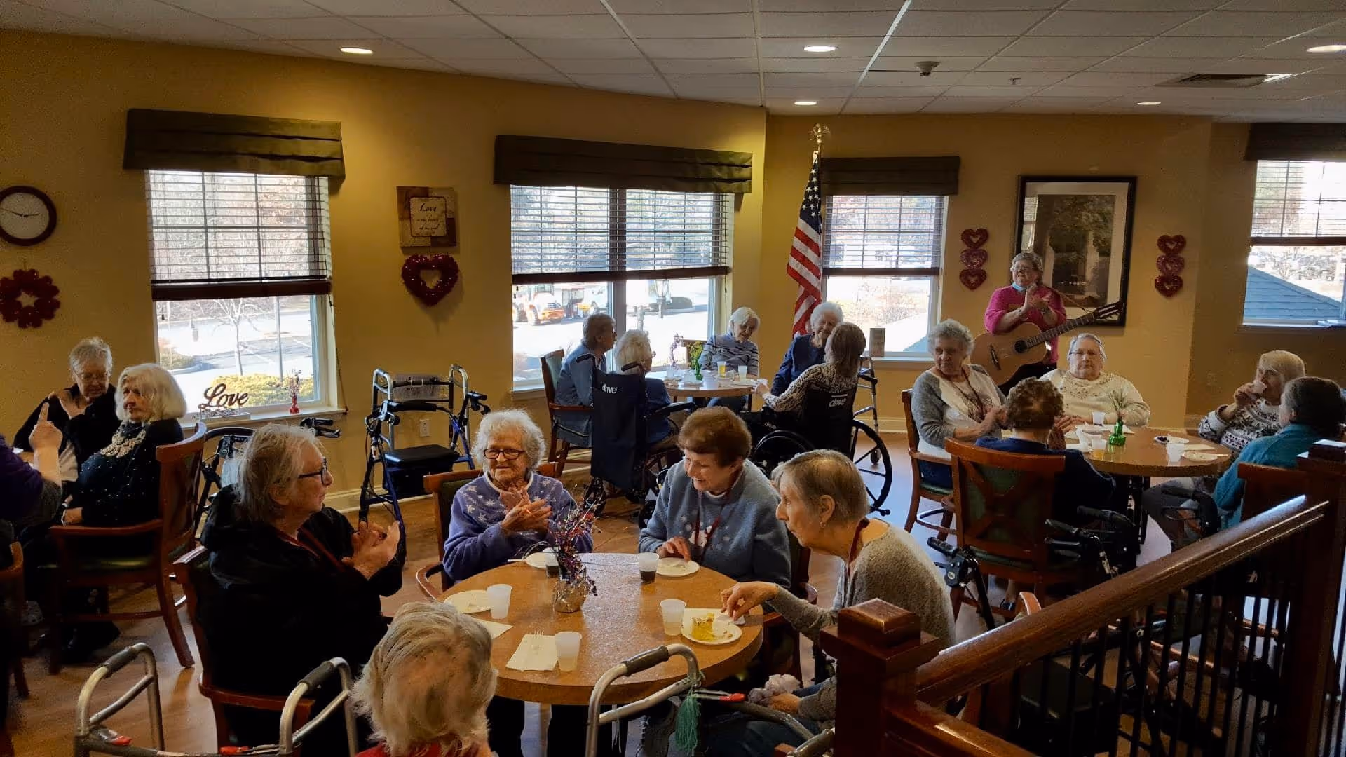 A group of elderly residents gathered at tables in a communal dining/activity room with a woman playing guitar by the windows and an American flag in the background.