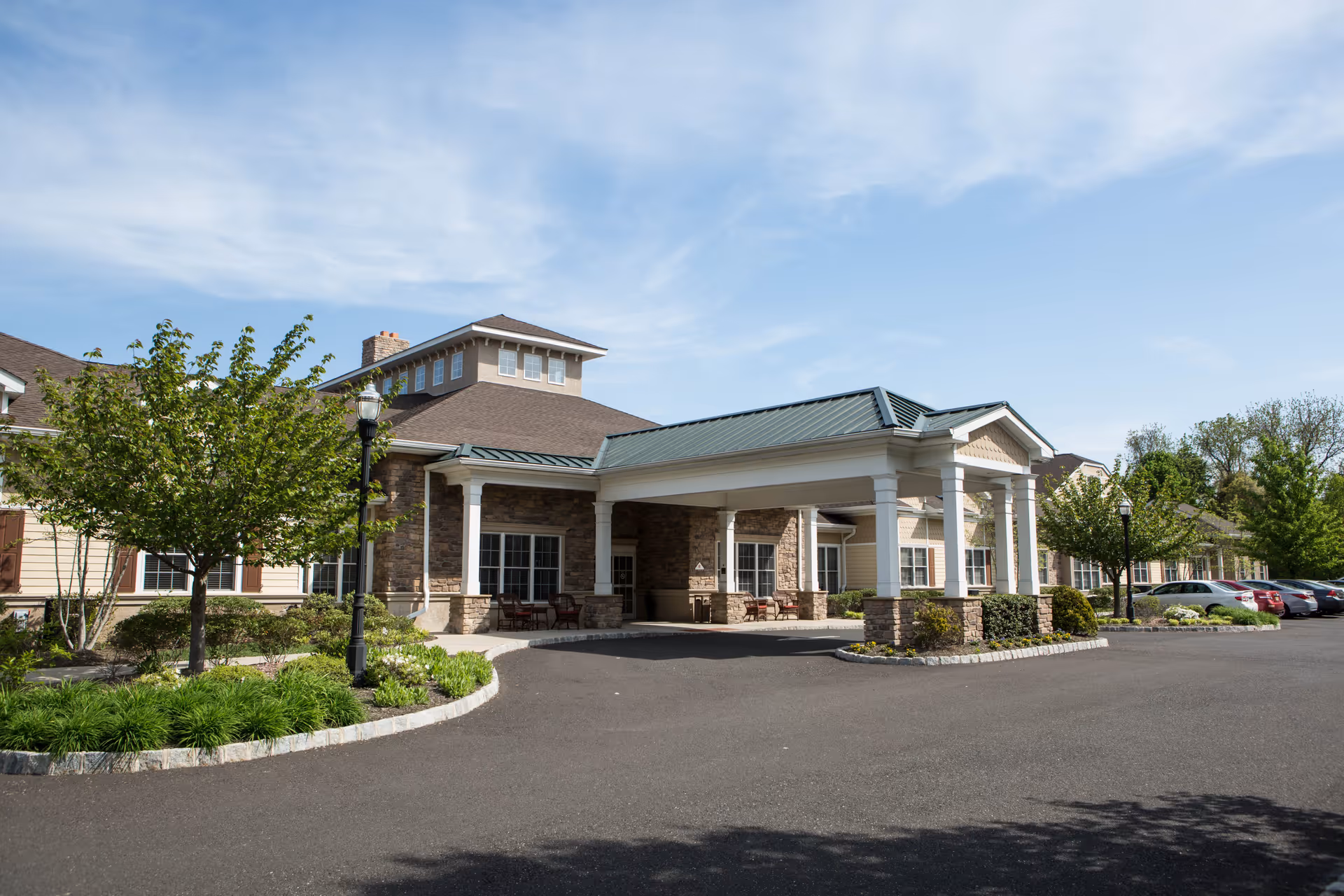 Exterior view of Harmony Village at Stanwick Road, showing the front entrance with a covered drop-off area supported by white columns. The building features a combination of stone and siding with multiple windows, surrounded by landscaped greenery and trees. Several cars are parked along the driveway under a clear blue sky.