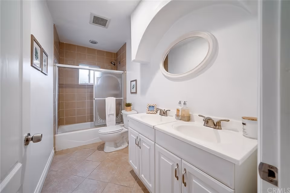 A clean bathroom with a white double sink vanity, an oval mirror above it, a toilet, and a bathtub with a sliding glass door and brown tiled walls. The floor is tiled, and there are small framed pictures on the walls.