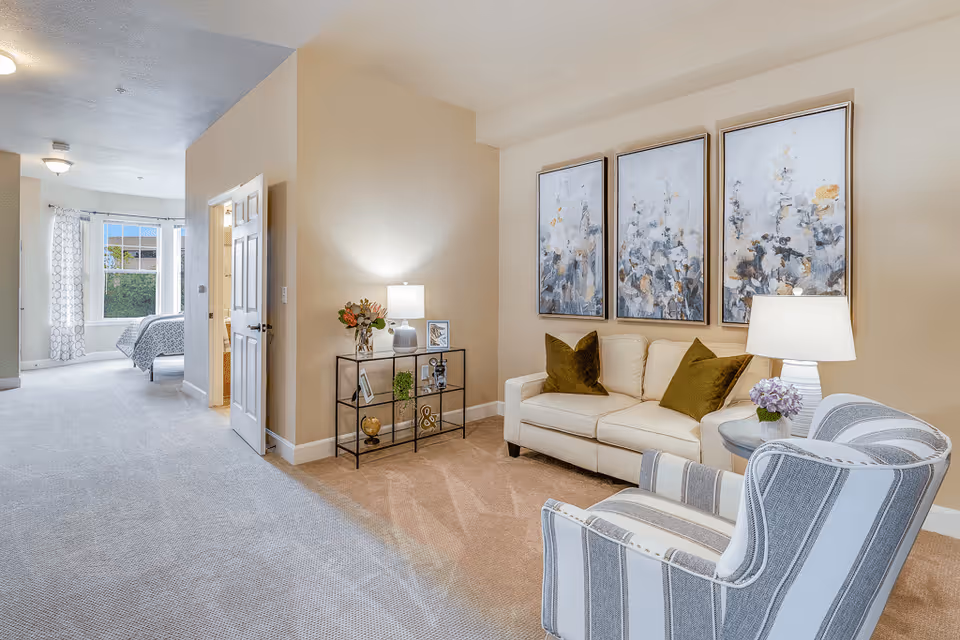 Interior living room with a sofa, striped armchair, side tables and three framed abstract floral paintings, opening into a bedroom.