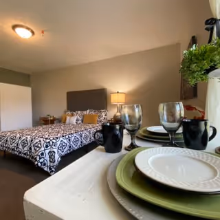 A cozy bedroom with a bed featuring a patterned black and white bedspread and decorative pillows. A bedside table with a lamp is next to the bed. In the foreground, a small dining table is set with plates, bowls, glasses, and mugs. A potted plant is visible near the window with curtains.