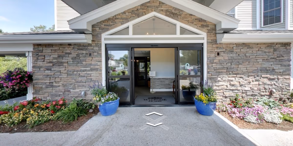 Entrance to a building with stone facade walls and a glass automatic sliding door. There are two large blue flower pots with colorful flowers on either side of the entrance, and flower beds with various plants and flowers along the sides. A welcome mat is visible inside the doorway.