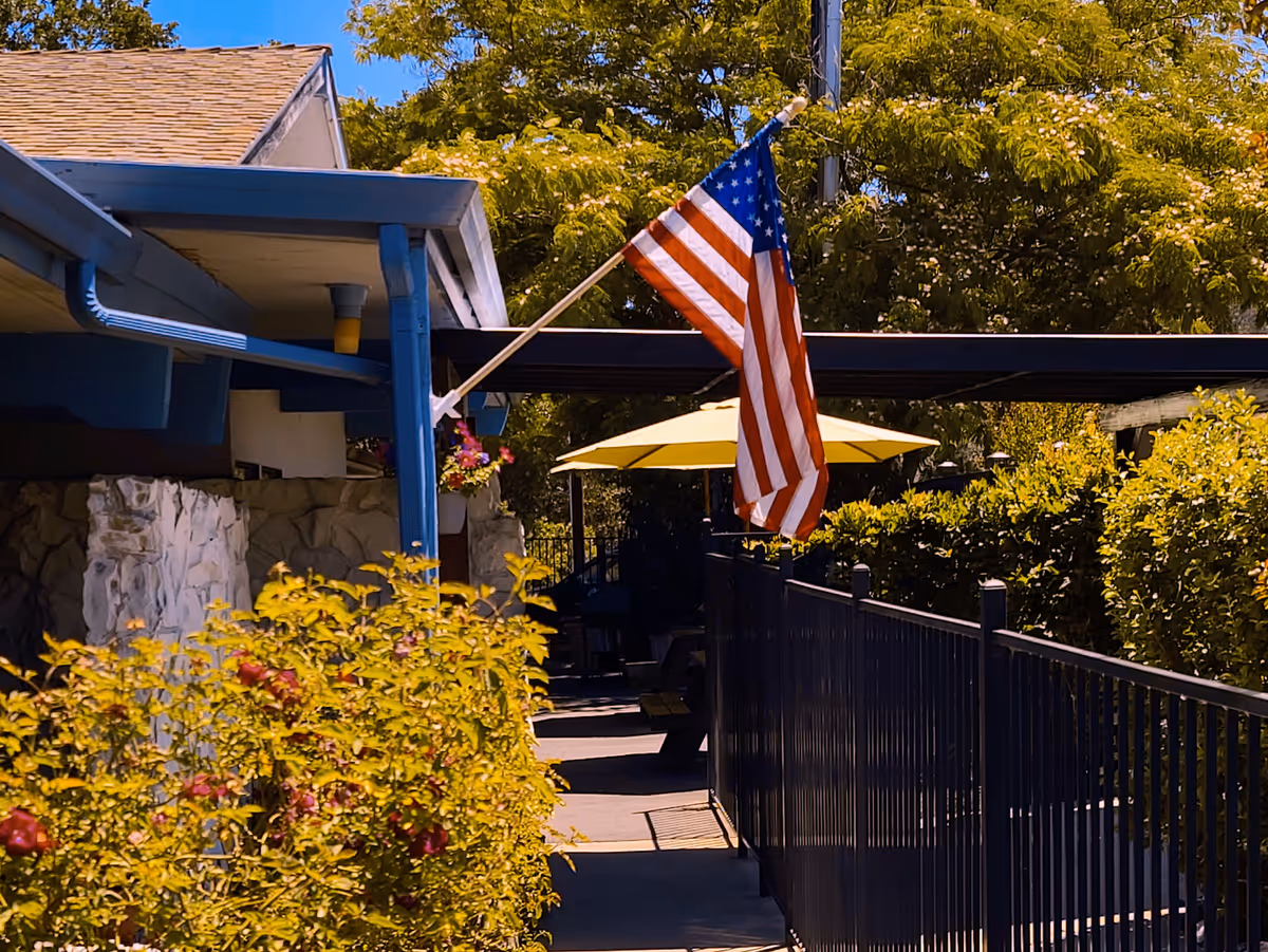 An outdoor walkway beside a senior living building with an American flag, a yellow patio umbrella, and surrounding greenery.