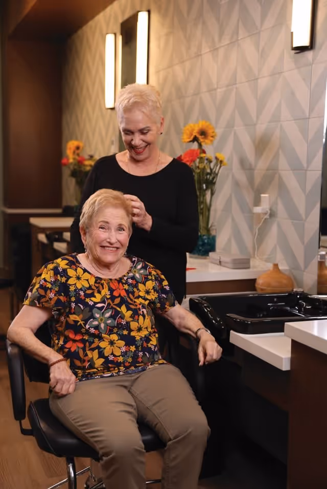 An elderly woman with a floral shirt sitting in a salon chair smiling while a hairstylist with short blonde hair stands behind her, styling her hair. The background shows a modern interior with geometric patterned walls, flowers in vases, and salon sinks.