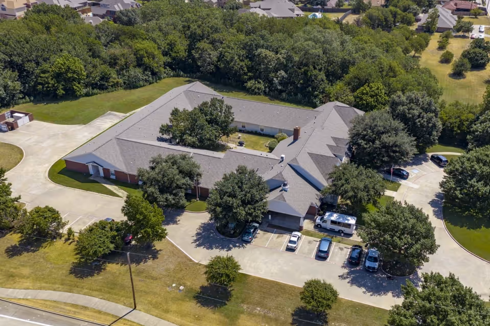 Aerial view of Deer Creek Senior Living facility showing a large single-story building with a central courtyard, surrounded by trees and parking spaces with several cars parked. The building is situated in a suburban area with other houses and greenery nearby.