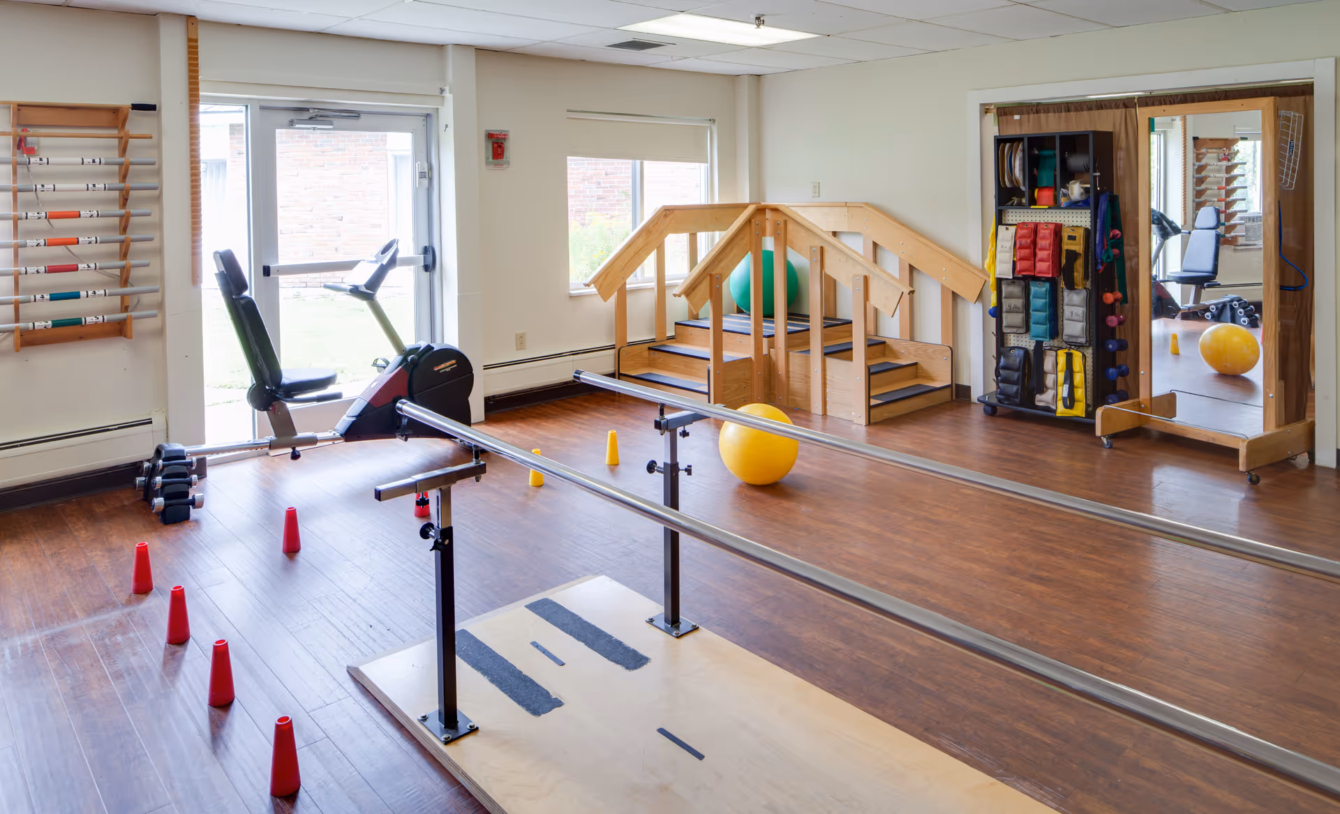 Physical therapy room with parallel bars for walking exercises, a stationary recumbent bike, small red and yellow cones on the floor, a wooden staircase with handrails, a large mirror, and a rack holding various exercise equipment including weights and balance pads.