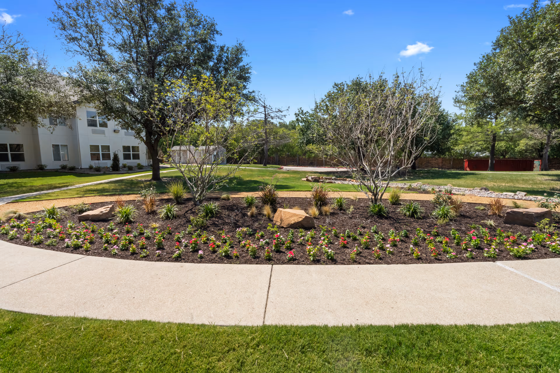 A landscaped garden area with a curved concrete pathway in front. The garden features small flowering plants, shrubs, and a few trees. In the background, there is a white building partially visible under a clear blue sky.