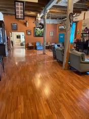 Spacious interior room with wooden flooring, exposed wooden beams on the ceiling, and orange walls. The room contains several seating areas with armchairs and sofas, framed pictures on the walls, and a ceiling fan. There is a doorway leading to another room in the background.