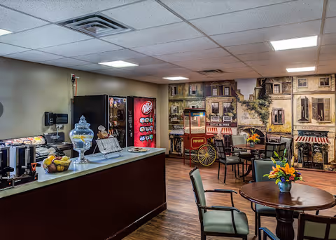 Interior of a senior living facility common area with a snack and beverage counter on the left, including a soda vending machine and a glass jar with snacks. Several round tables with chairs are arranged in the room, each table decorated with a small flower arrangement. The back wall features a mural depicting a quaint street scene with buildings and storefronts, and a vintage popcorn machine is positioned near the mural.