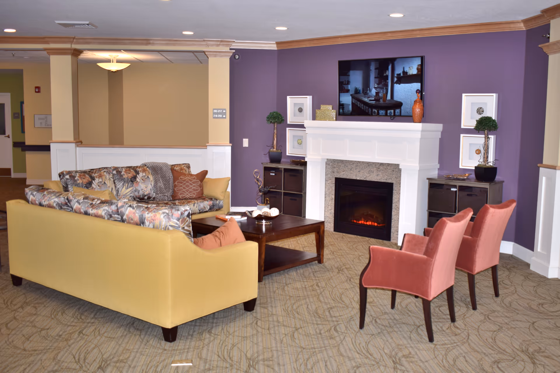 A cozy living room area in a senior living facility with a yellow sofa featuring floral cushions, two pink upholstered chairs, a wooden coffee table, and a white fireplace with a TV mounted above it on a purple accent wall. Decorative plants and framed artwork flank the fireplace.