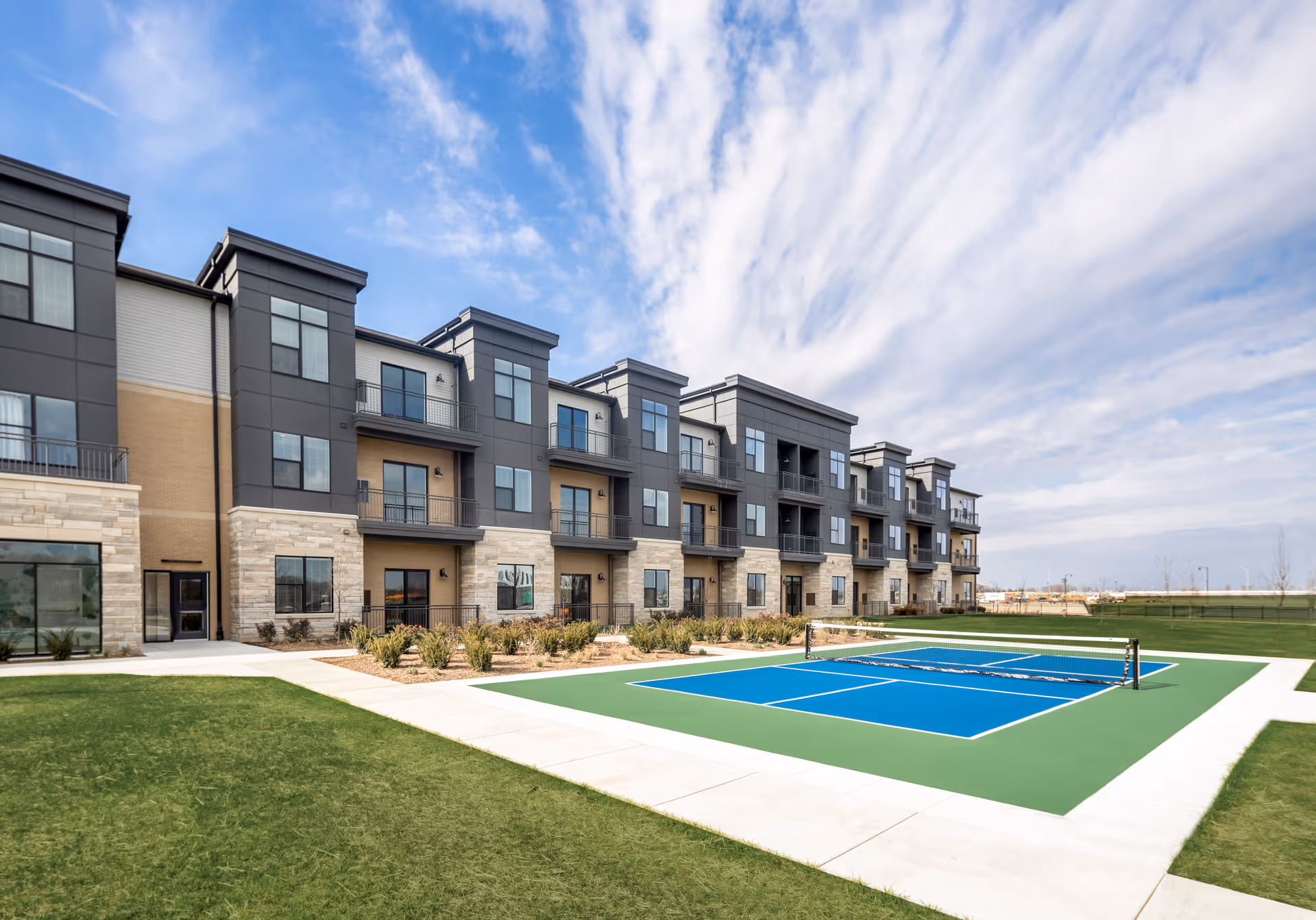 Modern three-story senior living building with balconies and an outdoor pickleball court on a sunny, partly cloudy day.