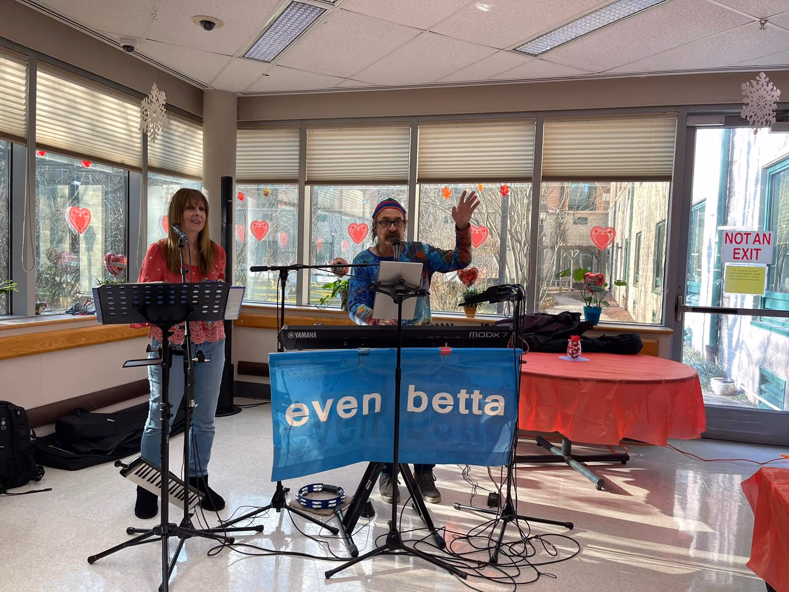 Two musicians performing indoors at Elmwood Hills Healthcare Center. One person is standing and singing into a microphone, while the other is playing a keyboard and waving. The room has large windows decorated with red heart-shaped stickers and snowflake decorations hanging from the ceiling. Tables covered with red tablecloths are visible in the background.