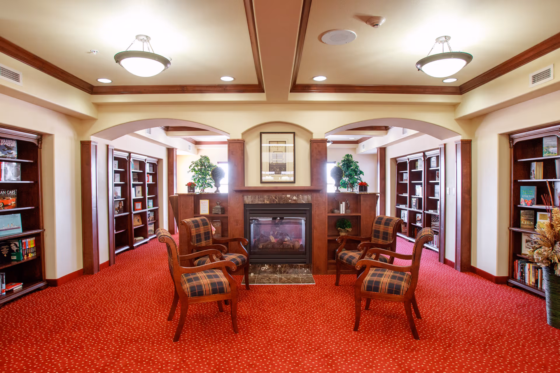 A cozy interior common area with red carpet and wooden trim. Four plaid upholstered chairs are arranged around a central fireplace with a marble base. Built-in wooden bookshelves line the walls on either side, filled with books and decorative items. Two potted plants are placed on either side of the fireplace, and framed artwork hangs above it. The ceiling features recessed lighting and two round ceiling lights.