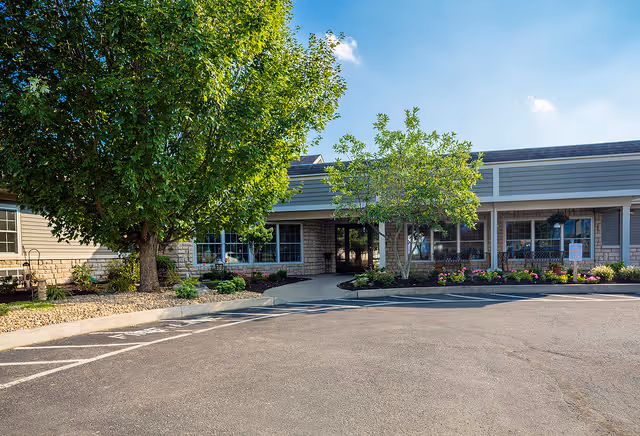 Front entrance of a single-story senior living facility with trees, landscaped flowerbeds, and a paved parking area.