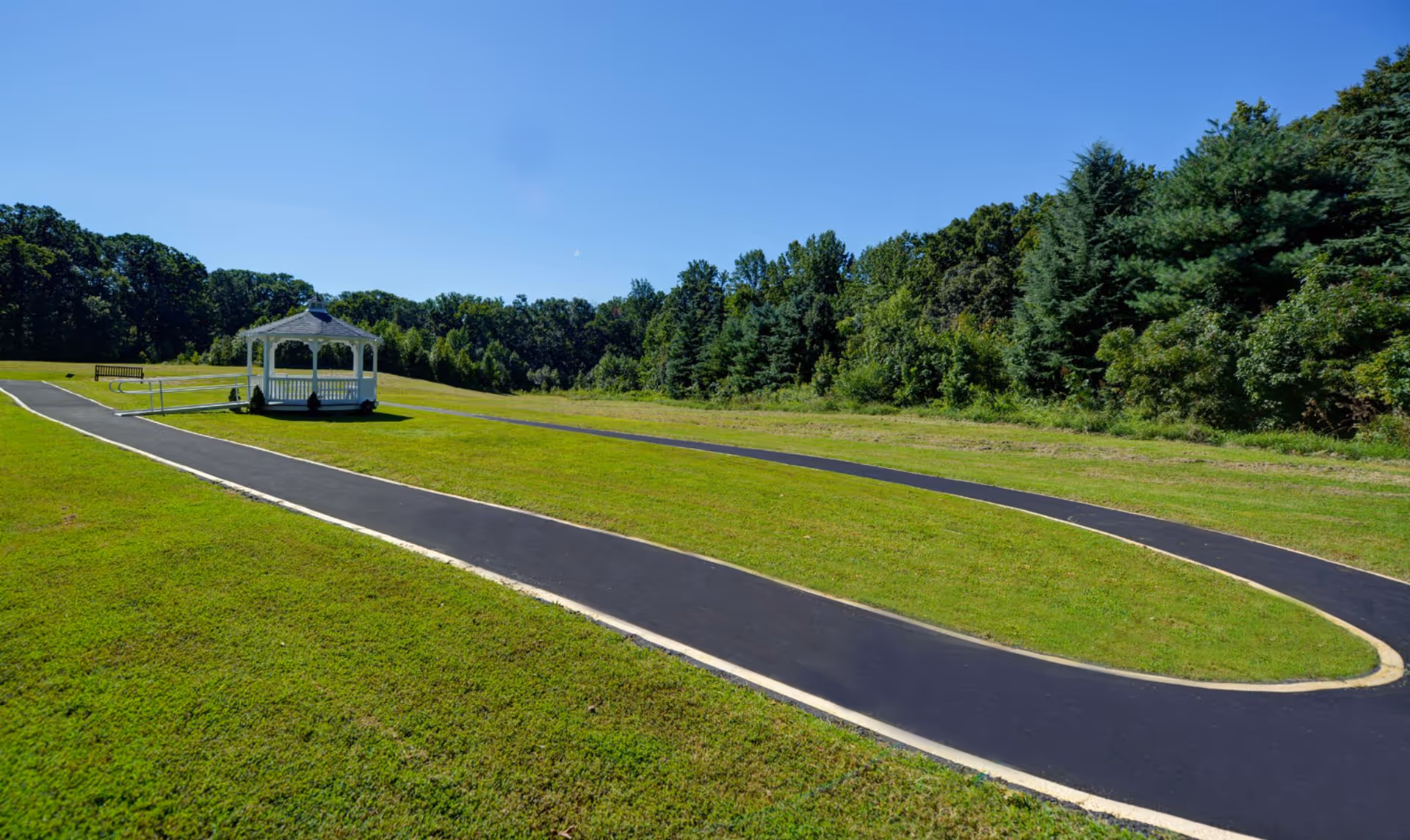 A paved walking path curves through a large grassy area with a white gazebo and a bench, surrounded by trees under a clear blue sky.