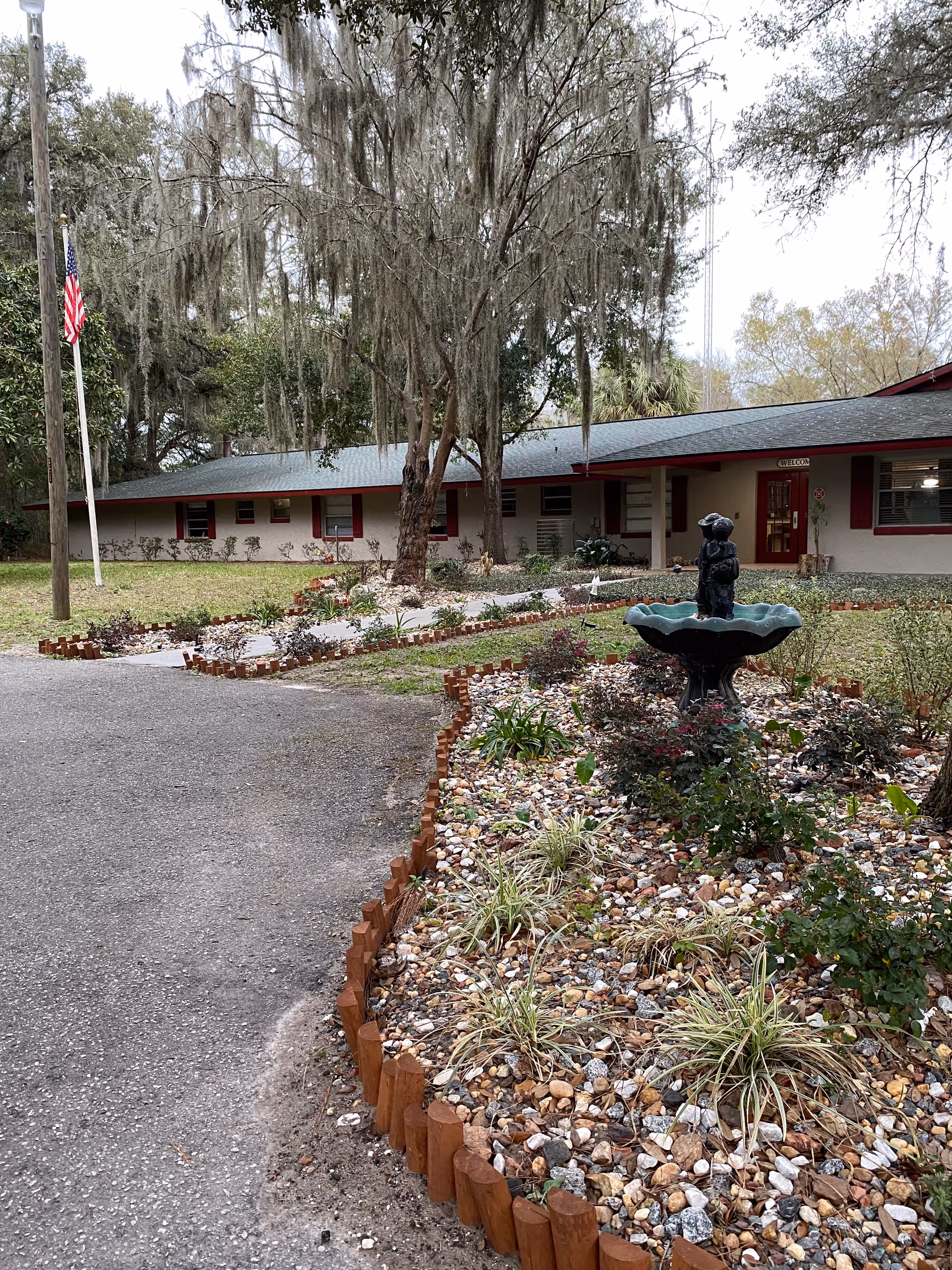 Exterior view of The Gardens Assisted Living Facility & Memory Care showing a single-story building with a gray roof and red window shutters. In front of the building is a landscaped garden area with a small fountain featuring a statue, surrounded by plants and decorative rocks. An American flag is visible on a flagpole to the left, and large trees with hanging moss provide shade.