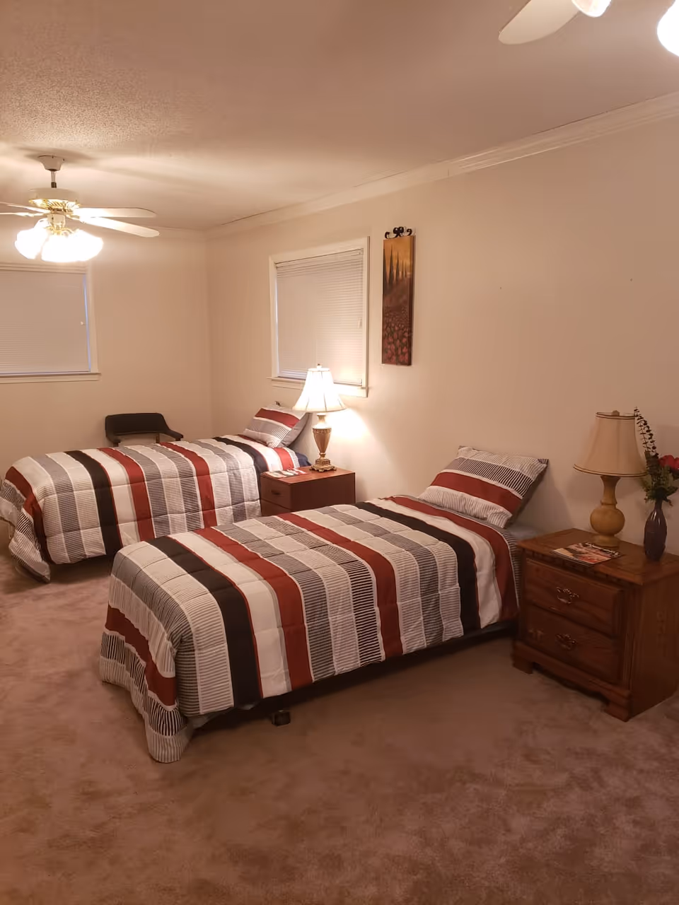 A bedroom with two single beds, each covered with striped bedspreads in shades of red, white, black, and gray. Between the beds is a small wooden nightstand with a lamp on it. Another wooden nightstand with a lamp and a vase with flowers is next to the bed on the right. The room has beige carpet, two windows with closed blinds, a ceiling fan with lights, and a wall decoration between the windows.