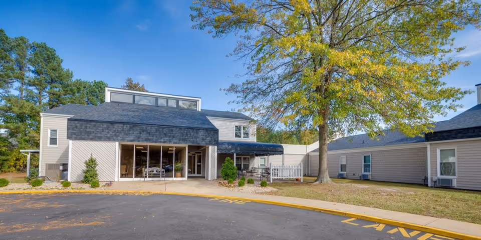 Exterior view of Parsons Residential Care Center showing a two-story building with large windows and an entrance canopy. The building is surrounded by a paved driveway, a large tree with green and yellow leaves, and some landscaping with bushes and grass under a clear blue sky.