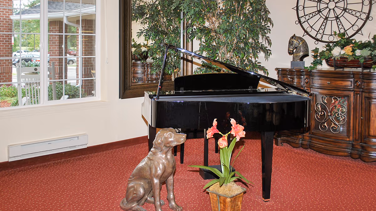 Black grand piano in a carpeted sitting area with a bronze dog statue, potted flowers, ornate sideboard and large window.