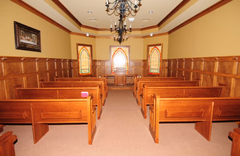 Interior view of a small chapel or meeting room with wooden pews arranged in rows facing a wooden podium. The room has yellow walls with wood paneling and three stained glass windows at the front. Two chandeliers hang from the ceiling.