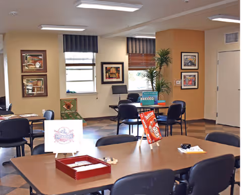 A well-lit common room with several tables and chairs arranged for group activities. On the tables are board games and cards, including a visible game of Yahtzee. The walls are decorated with framed artwork, and there are windows with blinds letting in natural light. A tall potted plant is placed near the corner of the room.