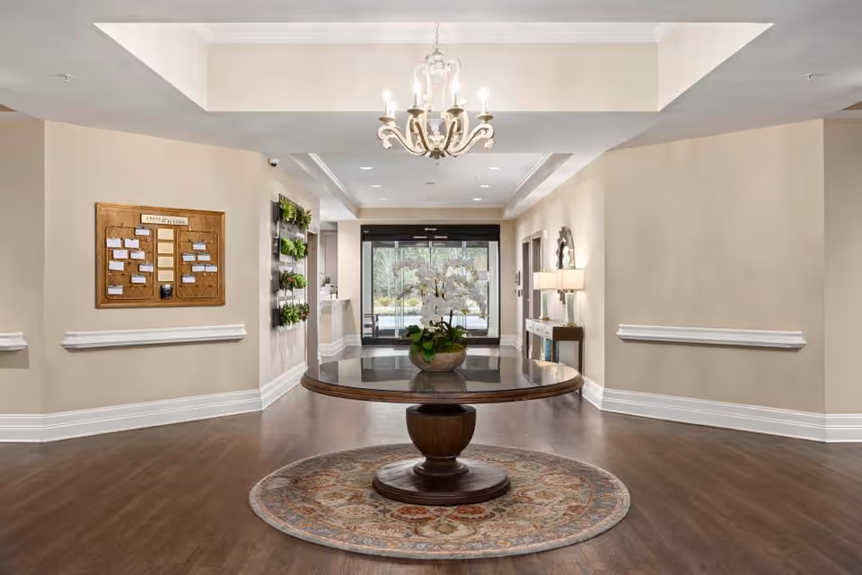 A spacious and well-lit lobby area with a round wooden table in the center holding a potted white orchid. The floor is wooden with a decorative round rug under the table. There is a chandelier hanging from the ceiling. On the left wall, there is a bulletin board and some wall-mounted plants. On the right side, there is a console table with a lamp and a mirror above it. Large glass doors at the far end lead outside.