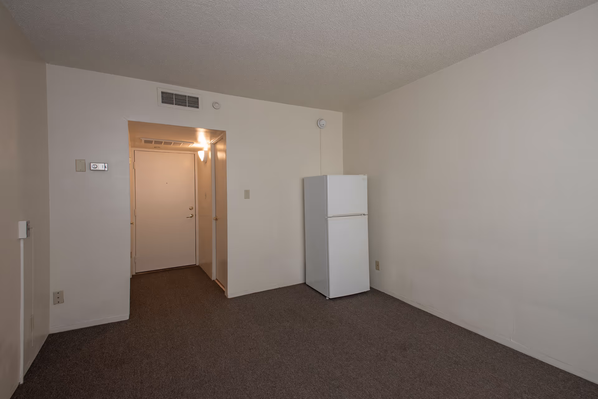 Empty room with beige walls and carpeted floor, a white refrigerator against the right wall, and a hallway leading to a closed door with a light fixture above it.