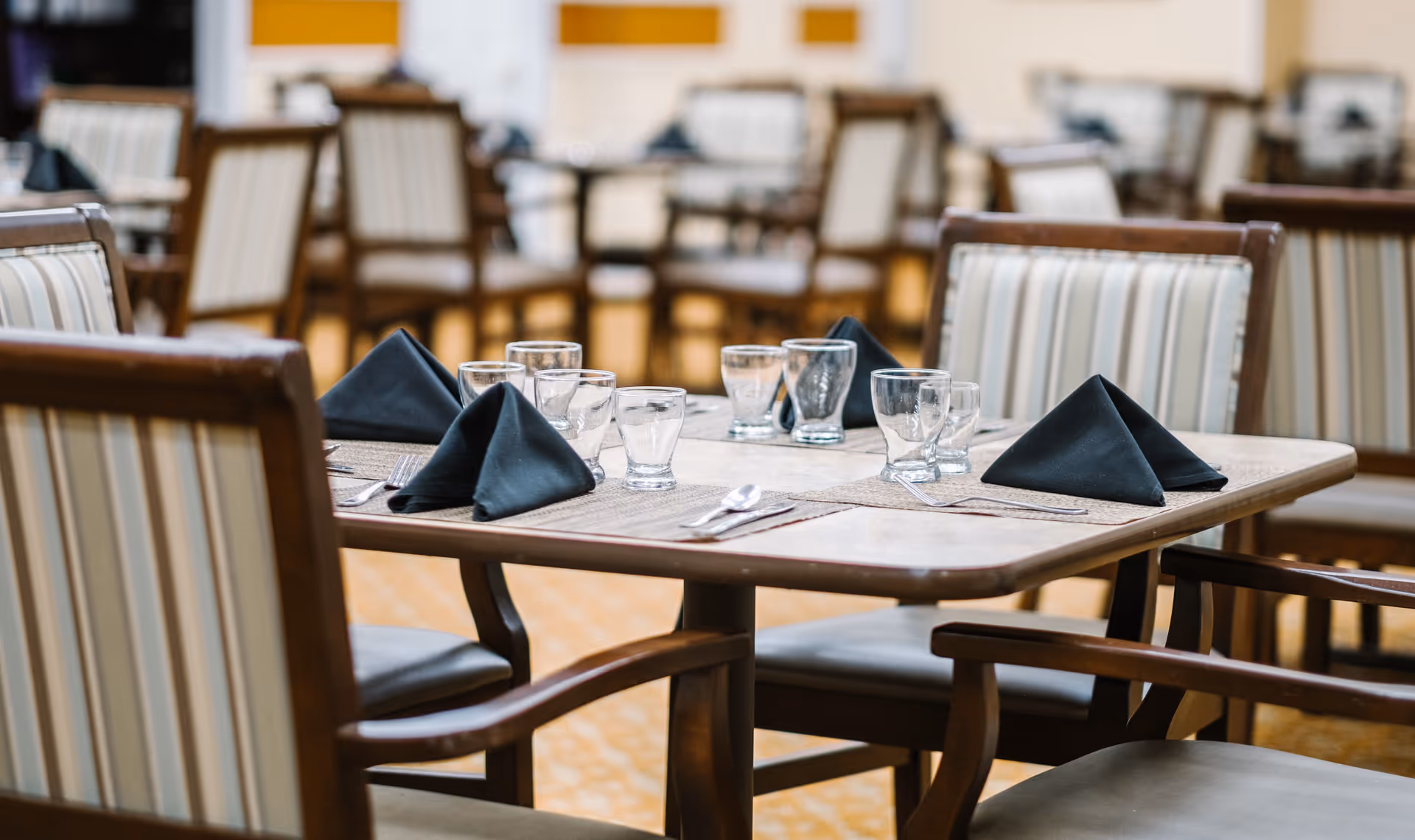 A dining table set with black folded napkins, clear drinking glasses, and silverware in a senior living facility dining room with striped cushioned chairs.