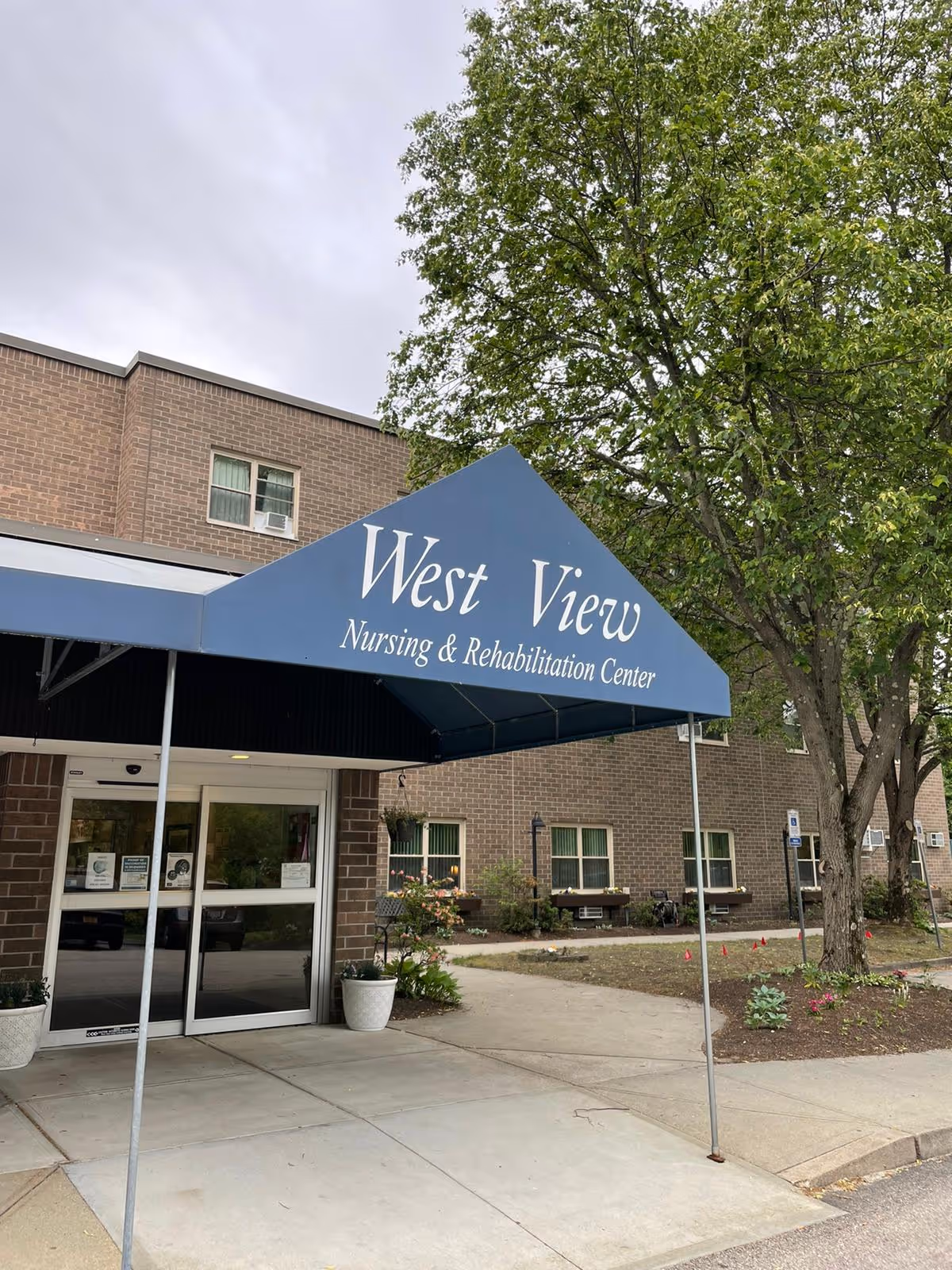 Entrance of West View Nursing & Rehabilitation Center with a blue awning displaying the facility's name. The building is made of brown brick, and there are trees and plants around the entrance area.