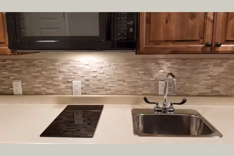 Kitchen countertop with a stainless steel sink and faucet, glass electric cooktop, microwave above, and a stone tile backsplash beneath wooden cabinets.