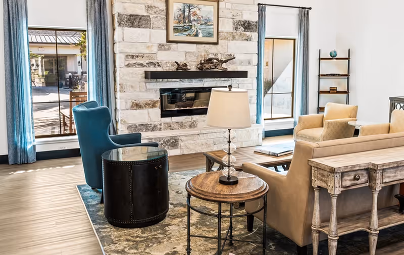 Bright communal living room with a stone fireplace, sofas and chairs, side tables, and large windows.