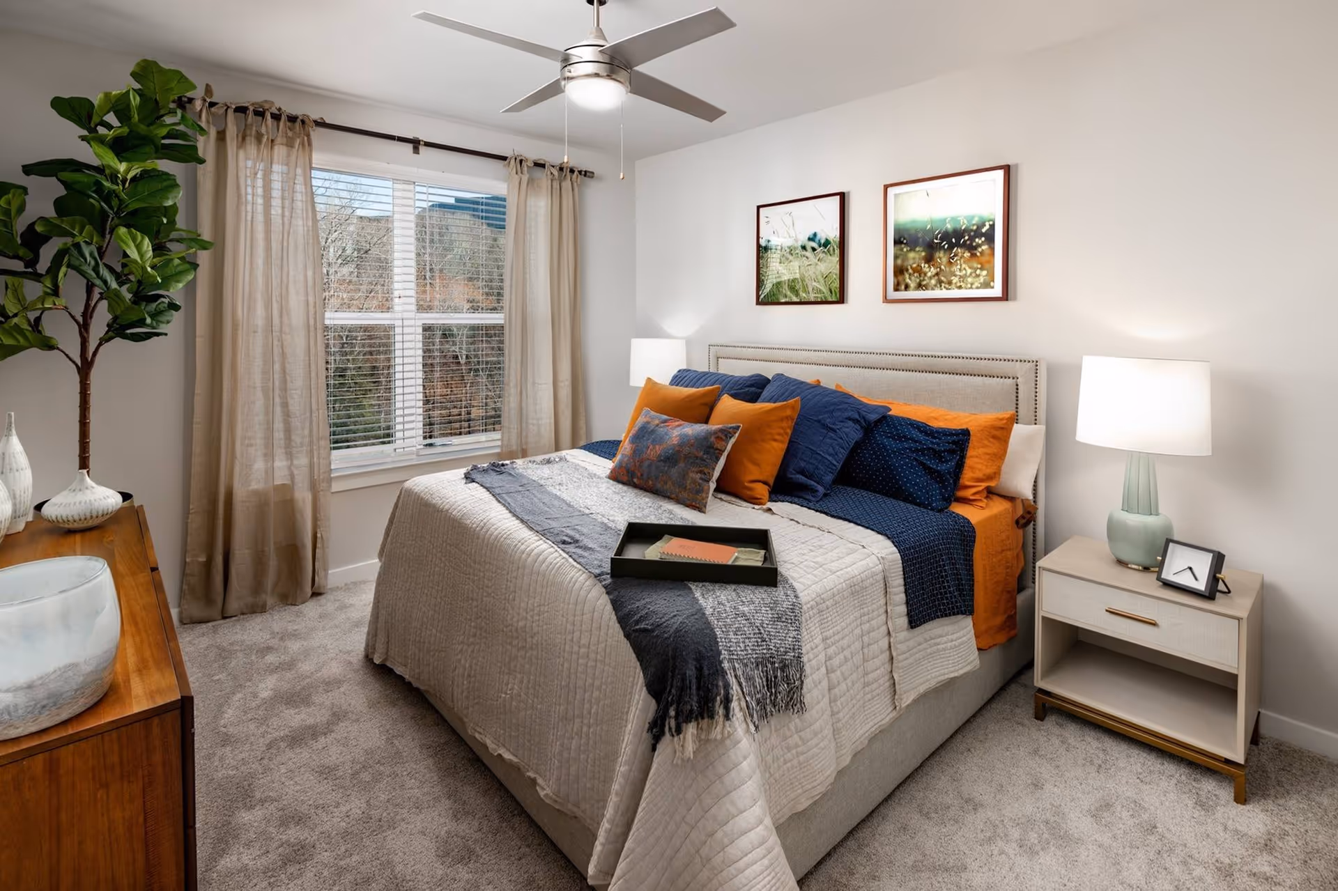 Well-lit furnished bedroom with a bed dressed in layered pillows and throws, a nightstand with lamp, a window with curtains, and a ceiling fan.