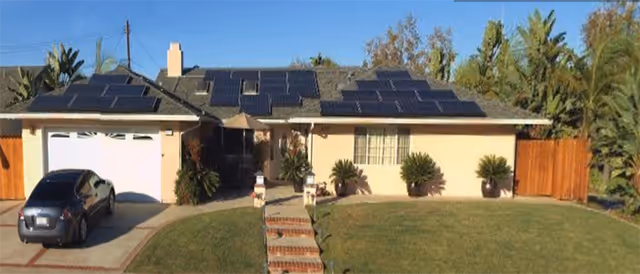 Single-story residential building with solar panels on the roof, a driveway with a parked car, a well-maintained lawn, and a brick walkway leading to the front door, surrounded by trees and shrubs under a clear blue sky.
