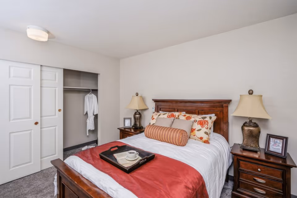 A neatly made bed with white and red bedding, decorative pillows, and a tray with a cup and napkin on top. The bed has a wooden headboard and is flanked by two wooden nightstands, each with a lamp and a framed photo. To the left, there is an open closet with sliding white doors and a white robe hanging inside. The room has light-colored walls and carpeted flooring.