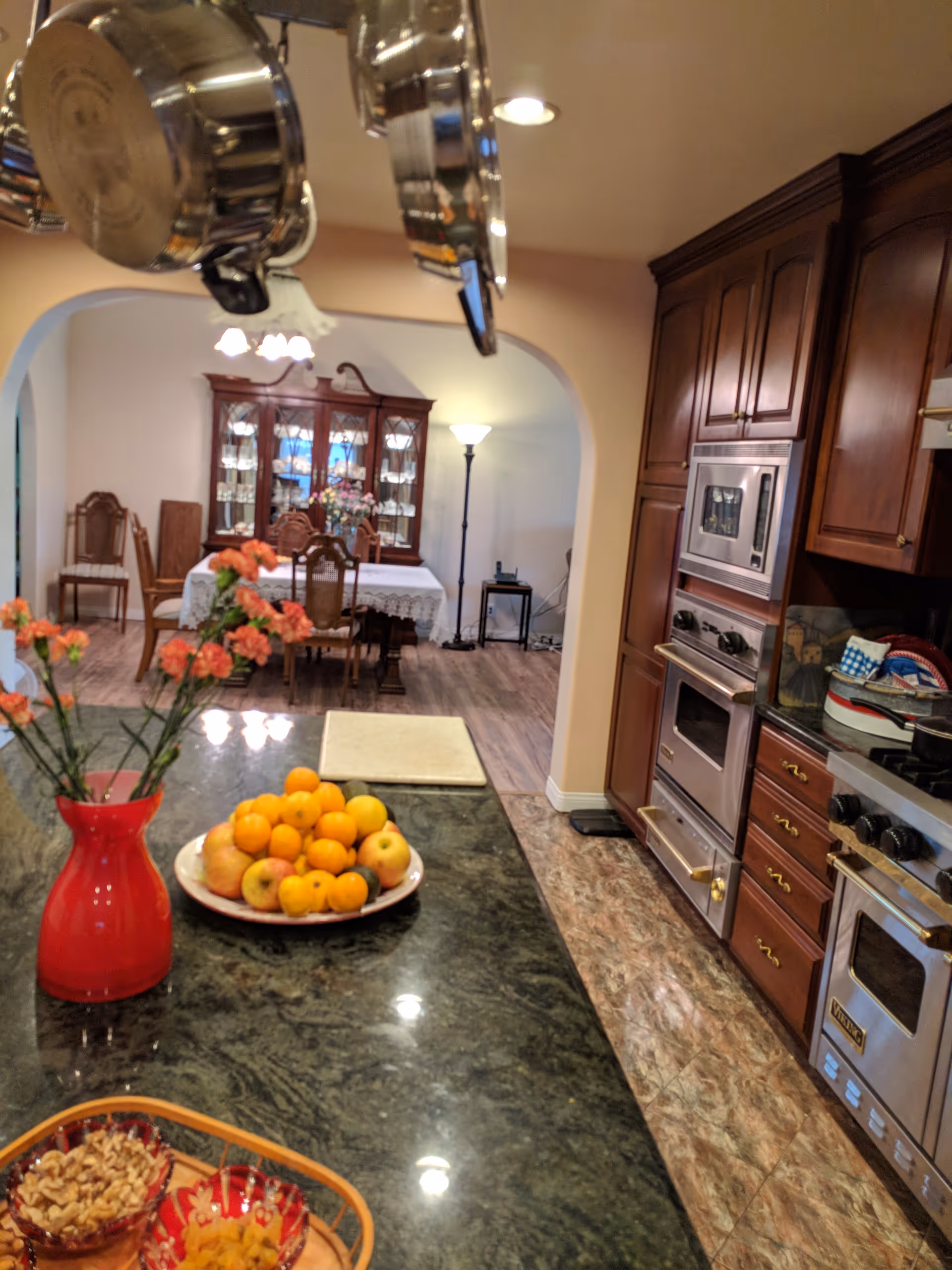 View of a kitchen with a dark granite countertop island in the foreground holding a red vase with orange flowers, a plate of assorted fruits including apples and oranges, and bowls with nuts and dried fruits. Stainless steel pots hang from the ceiling above the island. The kitchen has wooden cabinets and built-in stainless steel appliances. In the background, through an arched doorway, there is a dining room with a table covered with a white lace tablecloth, wooden chairs, a china cabinet, and a floor lamp.